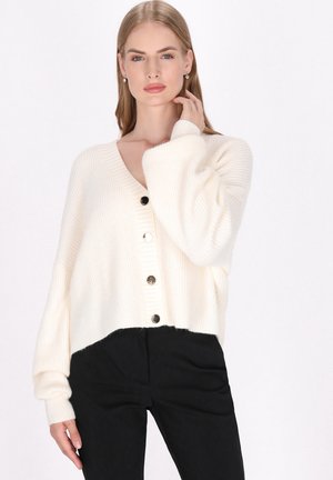 Vaalean kerma beige neulecardigan, jossa on V-pääntie, viisi mustaa nappia, putoavat olkapäät ja lyhyt malli. Pukeutunut tummien housujen päälle.