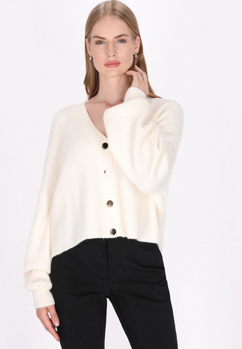 Cardigan tricotat de culoare crem deschis, cu decolteu în V, având cinci nasturi negri, umeri căzuți și un design scurt. Purtați peste pantaloni negri.