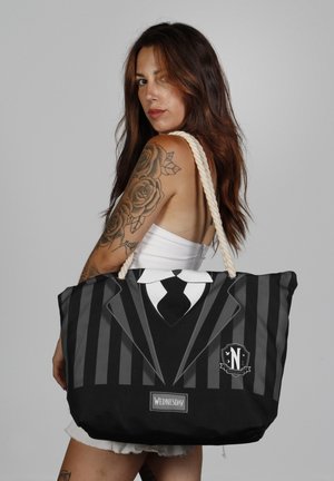 Bolsa tote negra diseñada para parecer un traje con rayas verticales blancas y grises; cuenta con un asa de cuerda y una etiqueta que dice "WEDNESDAY".