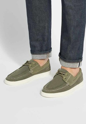 Baskets décontractées en mesh vert olive avec semelles blanches portées avec un jean en denim foncé retroussé sur un fond clair.