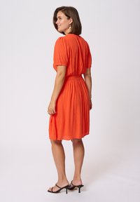 Robe orange à manches courtes avec taille froncée, forme A-line, tissu texturé et jupe plissée, associée à des sandales à talons noires.