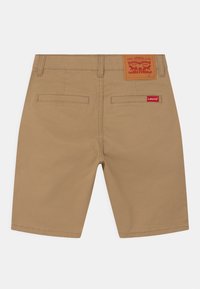 Béžové bavlněné šortky s zadními kapsami, koženou značkou a červeným logem. Hladká textura a rovný střih nohavic.