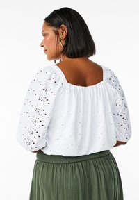 Blusa bianca in pizzo con maniche a palloncino e scollatura quadrata sul retro, abbinata a una gonna verde plissettata. Il design presenta delicate aperture.