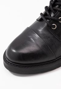 Zapato de cuero negro con puntera lisa y parte superior texturizada, que cuenta con cordones negros, ojetes plateados y una gruesa suela de goma negra.