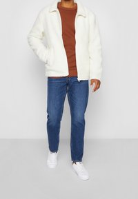 White fleece jacket with a zip front and collar, worn over a brown sweater, paired with blue jeans and white sneakers.