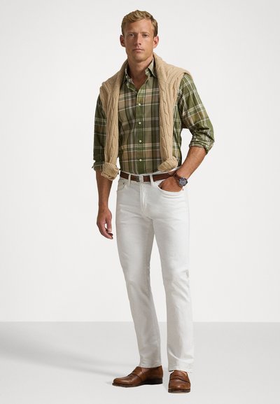 Πράσινο καρό πουκάμισο με μπεζ πλεκτή ζακέτα ριγμένη στους ώμους, λευκά παντελόνια και καφέ δερμάτινα παπούτσια. Ορατό απλό ρολόι χειρός.