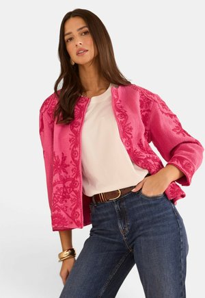 Donna con lunghi capelli castani indossa una giacca rosa acceso con motivo, top bianco, jeans scuri e cintura marrone, posando con una mano in tasca.