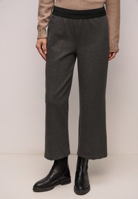 Person trägt dunkelgraue, strukturierte, weit geschnittene Hose, beigefarbenen Pullover und schwarze Leder-Stiefeletten, steht vor einem schlichten Hintergrund.