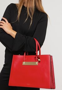 Bolso de cuero rojo con un diseño estructurado, dos asas y un acento de metal dorado. Textura suave con un acabado pulido y bordes en contraste.
