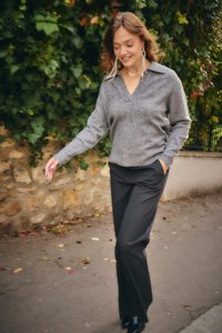 Ženska, ki hodi na prostem in se smehlja, oblečena v sivi pulover, črne hlače, dolge uhane in črne čevlje, z zelenim listnatim ozadjem.