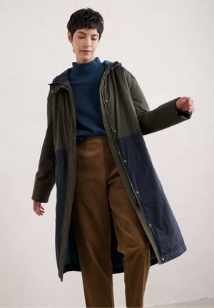 Lange, hoodie-jas in donkergroen en marineblauw, gemaakt van een lichtgewicht, glanzende stof en voorzien van drukknoopsluitingen. Gecombineerd met een bruine ribfluwelen broek.