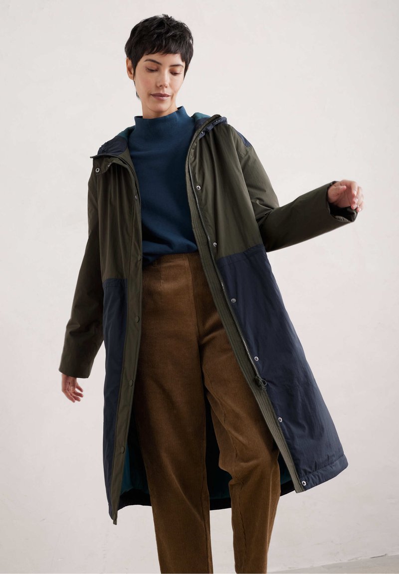 Lange, hoodie-jas in donkergroen en marineblauw, gemaakt van een lichtgewicht, glanzende stof en voorzien van drukknoopsluitingen. Gecombineerd met een bruine ribfluwelen broek.