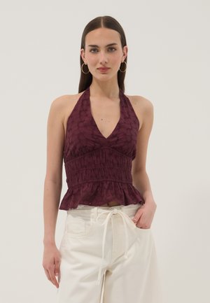 Mujer con top de cuello halter morado sin mangas con detalle de ojales y pantalones blancos de cintura alta, de pie con una mano en el bolsillo.