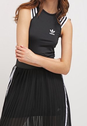Kobieta nosząca czarny bezrękawnik Adidas z białymi paskami i logo, zestawiony z czarną, plisowaną spódnicą z białymi bocznymi paskami.