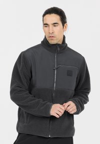 Tummanharmaa fleece-vuorattu takki, jossa on vetoketjullinen rintatasku, korkea kaulus ja yhdistelmä teksturoitua fleeceä sekä sileää kangasta.