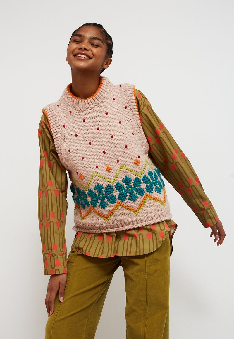 Persona sorridente che indossa un maglione senza maniche lavorato a maglia con motivi floreali sopra una camicia con motivi verdi e rossi e pantaloni color senape, posando.