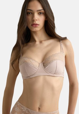 Ung kvinde med langt brunt hår iført lys pink blonde-bh og matchende undertøj mod en ensartet hvid baggrund.