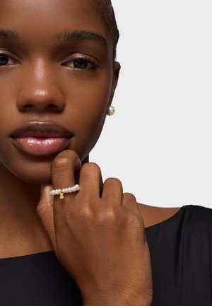 Anello con perla e accentuato in oro su una mano, che presenta perle bianche e una tonalità di pelle texturizzata. Sullo sfondo un grigio chiaro, con indumenti neri visibili.