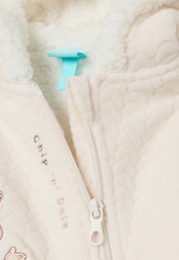 Chaqueta de cremallera de crema clara, hecha de una tela texturizada y suave. Presenta un forro suave, texto bordado y una etiqueta de tela turquesa.