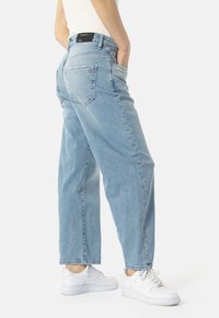 Osoba v světle modrých rovně střižených džínách s černou nášivkou na pase a bílými teniskami, stojící s rukama v kapsách.