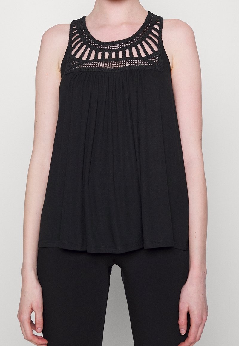 Haut noir sans manches avec un col en crochet, tissu plissé et coupe ample, associé à un pantalon noir ajusté.