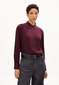 Long-sleeved, collared shirt in deep burgundy fabric, paired with high-waisted dark denim jeans and a black belt with a gold buckle.