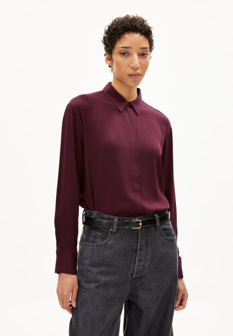 Long-sleeved, collared shirt in deep burgundy fabric, paired with high-waisted dark denim jeans and a black belt with a gold buckle.