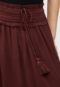 Høytlivs, burgunder skjørt med rynket, volangbelagt midjebånd knyttet med lange duskestrenger, brukt sammen med en svart topp.