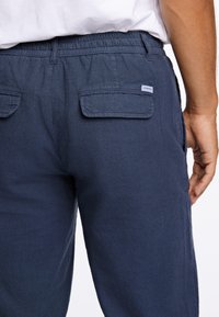 Navyblauwe linnen broek met een elastische tailleband, twee achterzakken en een subtiele textuur; etiket zichtbaar op de rechterzak.