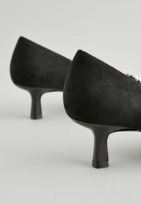 Svarta mockaskor med hög klack, spetsig tå och smal klack. Materialet har en slät, matt yta. Inga synliga detaljer eller mönster.