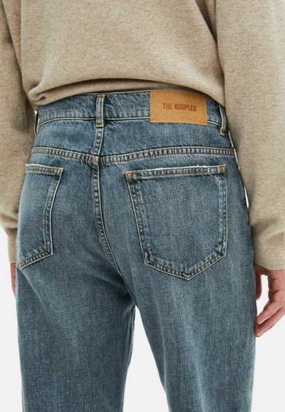 Jeans de mezclilla azul deslavado con un parche de cuero marrón etiquetado "THE KOOPLES", con bolsillos traseros estándar y detalles de costura visibles.