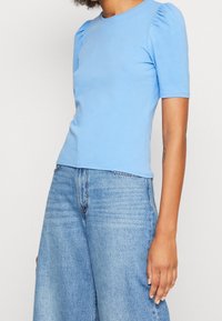 Světlomodrý krátký rukáv top s nabíranými rameny, hladkou texturou a kulatým výstřihem, doplněný volnými, jemně vybledlými modrými džíny.