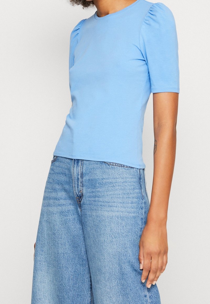 Světlomodrý krátký rukáv top s nabíranými rameny, hladkou texturou a kulatým výstřihem, doplněný volnými, jemně vybledlými modrými džíny.