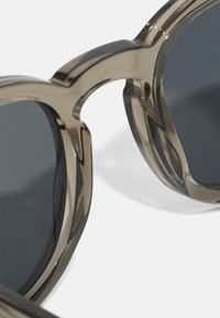 Sonnenbrille mit einer großen, runden Form, klarem braunem Kunststoffrahmen, dunkelgrauen Gläsern und skulpturalen Brückendetails.