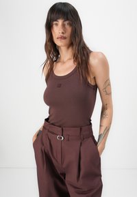 Brown ribbed tank top with a round neckline, features small logo detail. Paired with high-waisted brown trousers with a silver hardware accent.