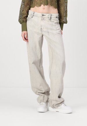 Personne portant un jean gris clair coupe ample, des baskets blanches et un chemisier court en dentelle vert olive à manches longues.