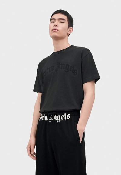 Νέος άνδρας που φοράει μαύρο t-shirt Palm Angels και μαύρο παντελόνι με το λογότυπο Palm Angels στη ζώνη, στέκεται με το ένα χέρι στην τσέπη.