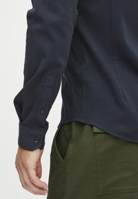 Cămașă albastru închis cu mâneci lungi și manșete cu buton, având o textură netedă și un tiv curbat. Asortată cu pantaloni verzi măslin cu buzunare laterale.
