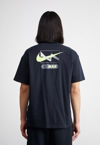 Schwarzes Baumwoll-T-Shirt mit kurzen Ärmeln. Auf der Rückseite befindet sich ein großes gelbes Nike-Logo und eine "AIR MAX" Grafik. Einfaches Design, lässiger Schnitt.