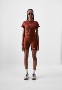 Brūns īss krekls ar logo, atbilstošas brūnas riteņbraukšanas šortus un sporta apavus. Modelis valkā melnas saulesbrilles un baltas zeķes līdz potītei.