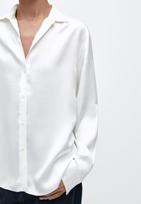 Camicia bianca a maniche lunghe con colletto standard e chiusura con bottoni, caratterizzata da una texture liscia e lucida e una vestibilità comoda.