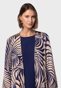 Satin kimonojacka med en tan bakgrund och ett marinblått botaniskt bladmönster, har en avslappnad passform och öppen framsida.