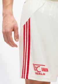 Κοντινό πλάνο ενός χεριού δίπλα σε λευκά αθλητικά σορτς με κόκκινες ρίγες και το λογότυπο "1. FC Union Berlin" στο αριστερό μηρό.