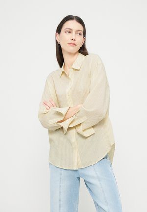 Γυναίκα με μακριά σκούρα μαλλιά που φοράει ένα ανοιχτό κίτρινο πουκάμισο με ρίγες και ανοιχτό μπλε τζιν, στεκόμενη με σταυρωμένα χέρια, κοιτάζοντας πλάγια.