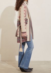 Mujer vestida con un cárdigan beige estampado con cinturón, jeans acampanados azules y botas de cuña negras con punta, de pie de lado.