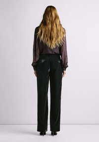 Pikkade blondide juustega naine, kes kannab musti kõrge vöökohaga sametpükse, tumedate triipudega pluusi ja musti kontsa. Ta seisab seljaga.