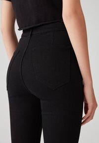 Jeans in denim nero a vita alta con vestibilità slim, dotati di due tasche posteriori e una texture liscia, mostrati da un'angolazione posteriore.