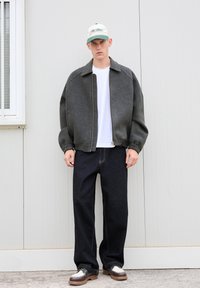 Giacca grigia, oversize, con colletto alto, indossata sopra una maglietta bianca. Pantaloni in denim scuro a gamba dritta e scarpe bicolore. Berretto con dettagli verdi.