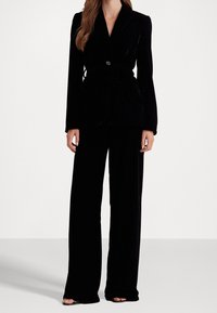 Costume en velours noir comprenant un blazer ajusté avec une taille ceinturée et un pantalon à jambes larges. La texture est douce et lisse, créant une silhouette élégante.