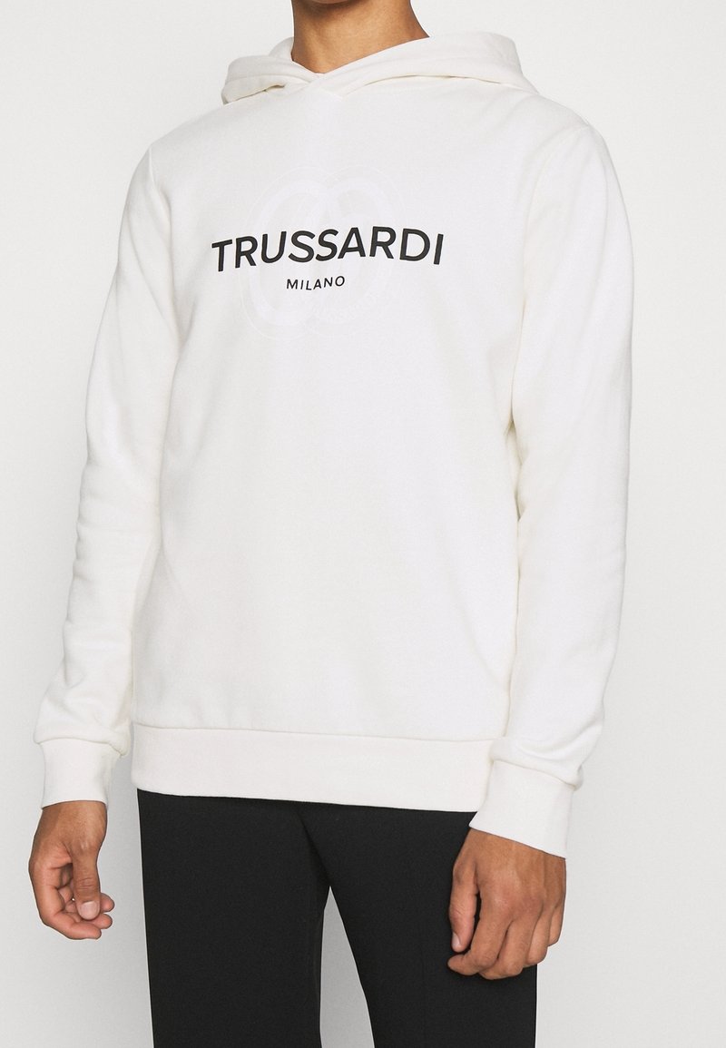 Personne portant un sweat à capuche blanc avec « TRUSSARDI MILANO » imprimé en noir sur le devant et un pantalon noir.